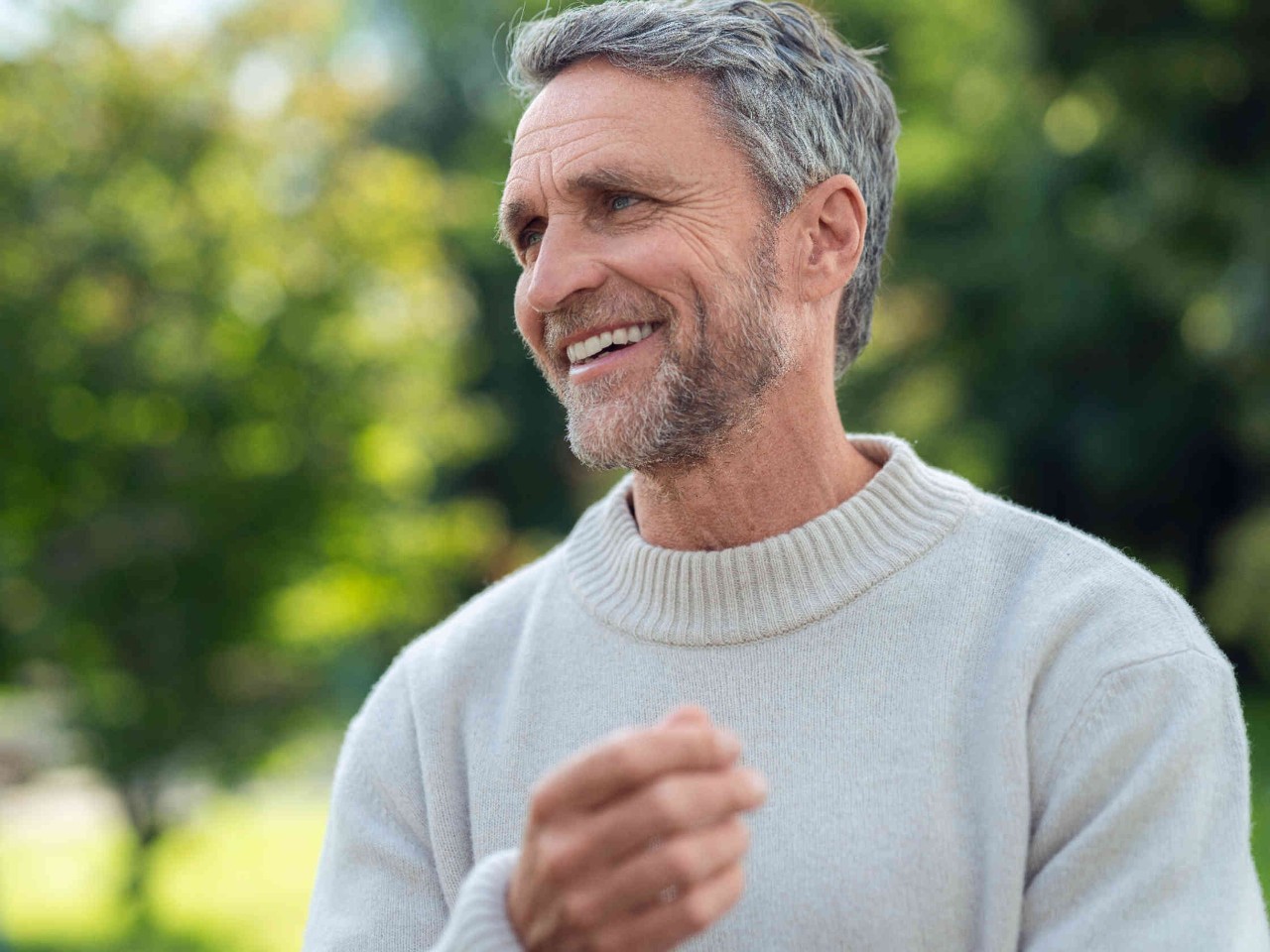 Mann mit grauem Haar und hellem Pullover steht im Freien und laechelt.