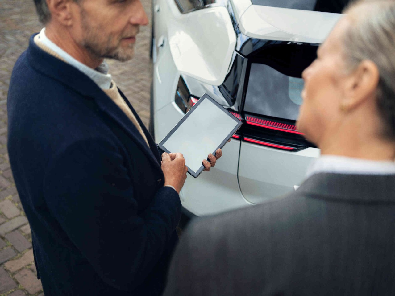 Mann haelt Tablet und steht neben Frau hinter einem weissen Auto auf Pflastersteinen.