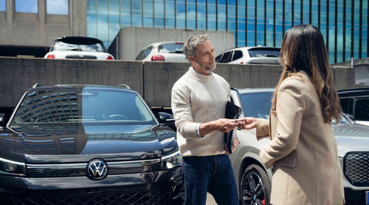 Mann ueberreicht Autoschluessel an Frau auf Parkplatz vor Buerogebaeude.