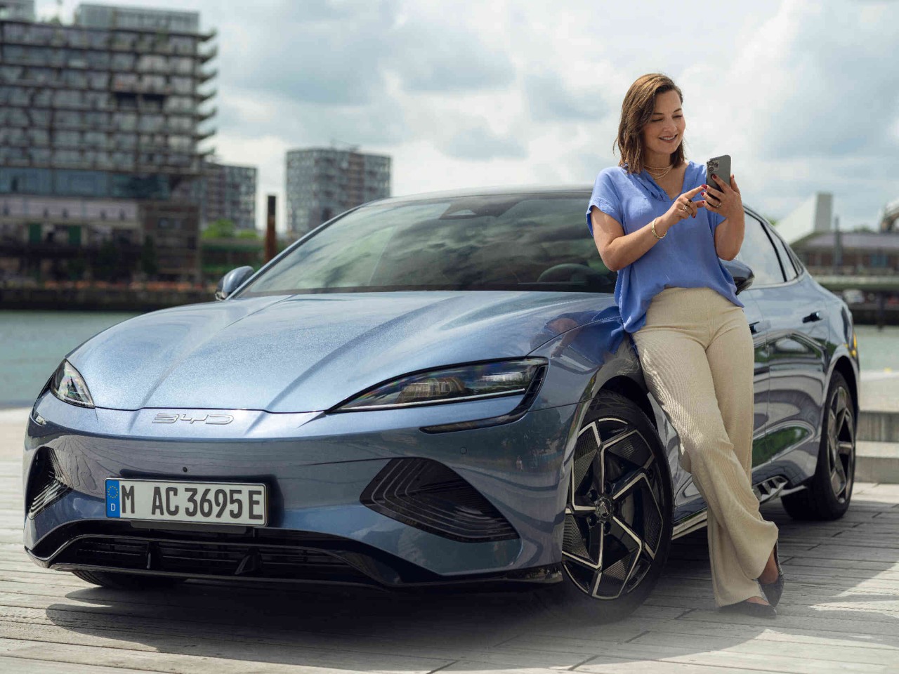 Eine Frau lehnt sich entspannt an ein blaugraues BYD Auto und nutzt ihr Handy w?hrend im Hintergrund ein Fluss und B?rogeb?ude zu sehen sind.