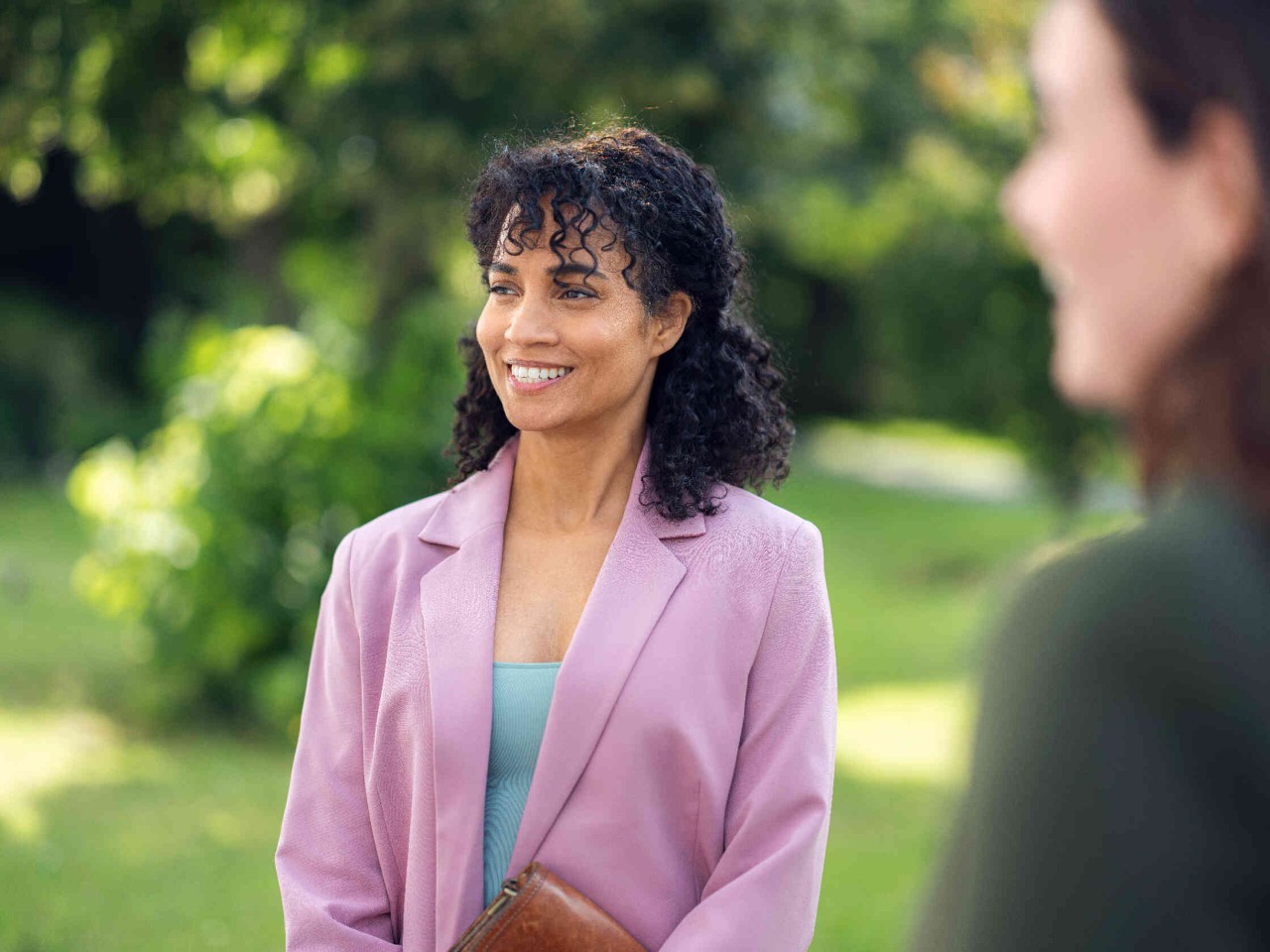 Frau in rosa Blazer und brauner Tasche steht in gruenem Park und laechelt.