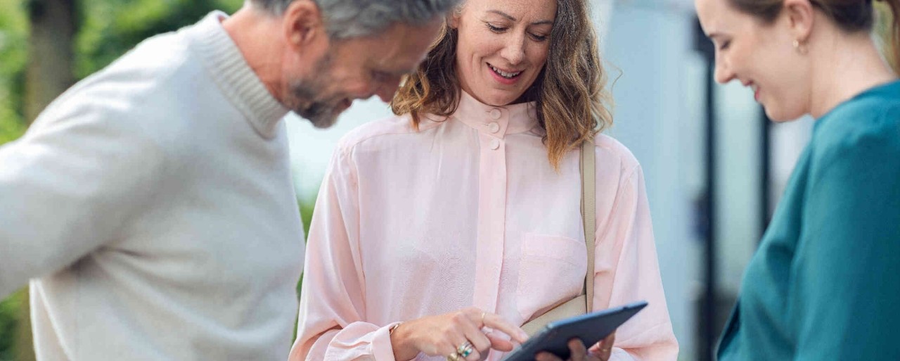 Frau haelt Tablet und blickt mit zwei weiteren Personen darauf.