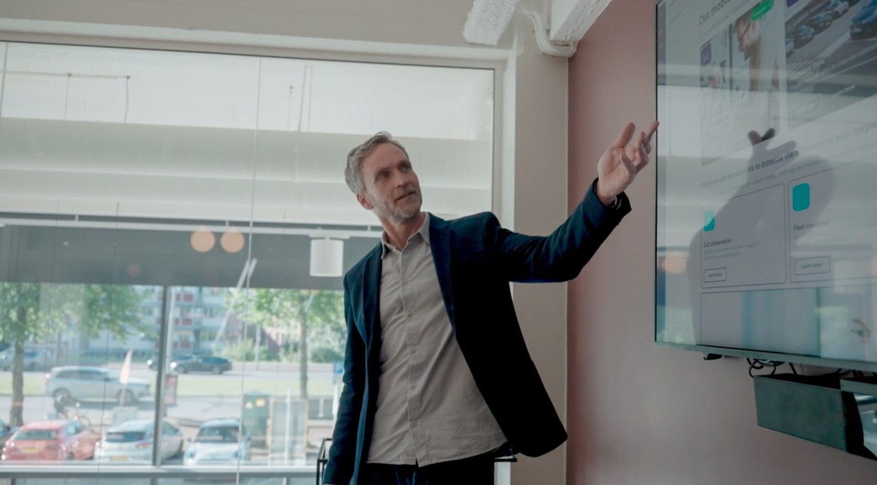 Ein Geschäftsmann hält vor Kollegen in einem modernen Konferenzraum eine Präsentation und zeigt dabei auf einer großen Leinwand die Emissionsdaten einer Fahrzeugflotte.