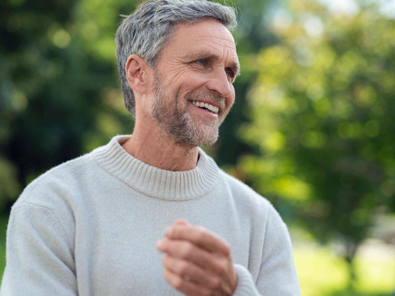 Mann mit grauem Haar und hellem Pullover steht im Freien und laechelt.