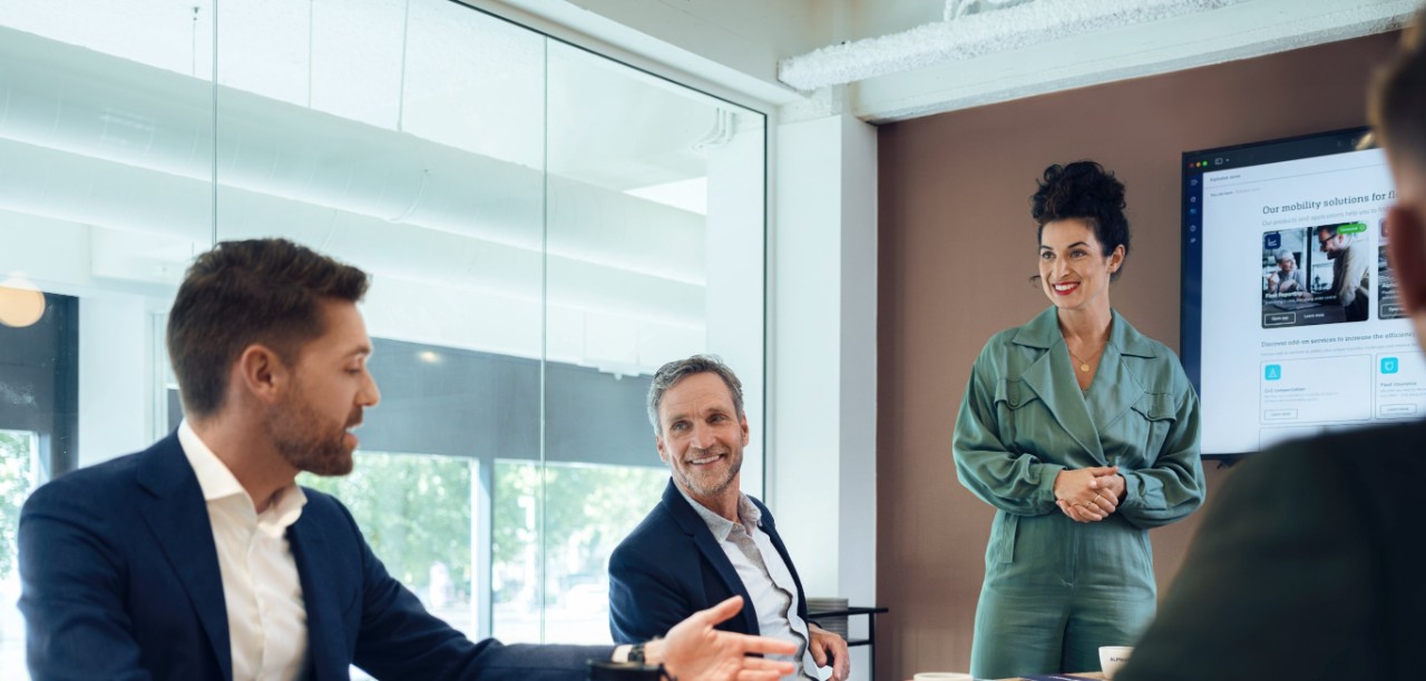 Drei Personen unterhalten sich an einem Holztisch in einem modernen Konferenzraum mit einem großen Bildschirm.
