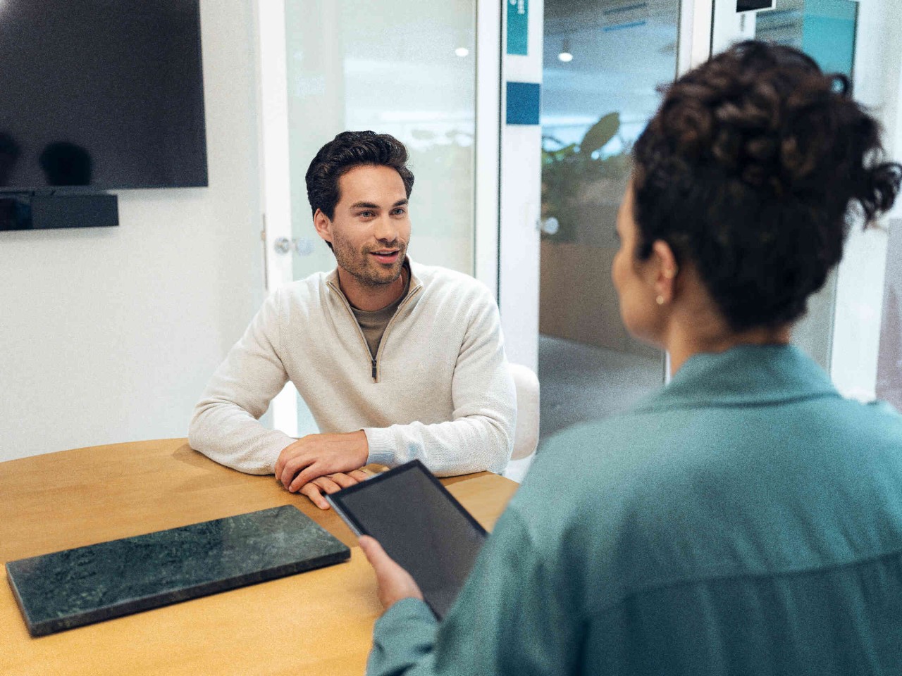 Frau mit Tablet im Gespraech mit Mann am Tisch.