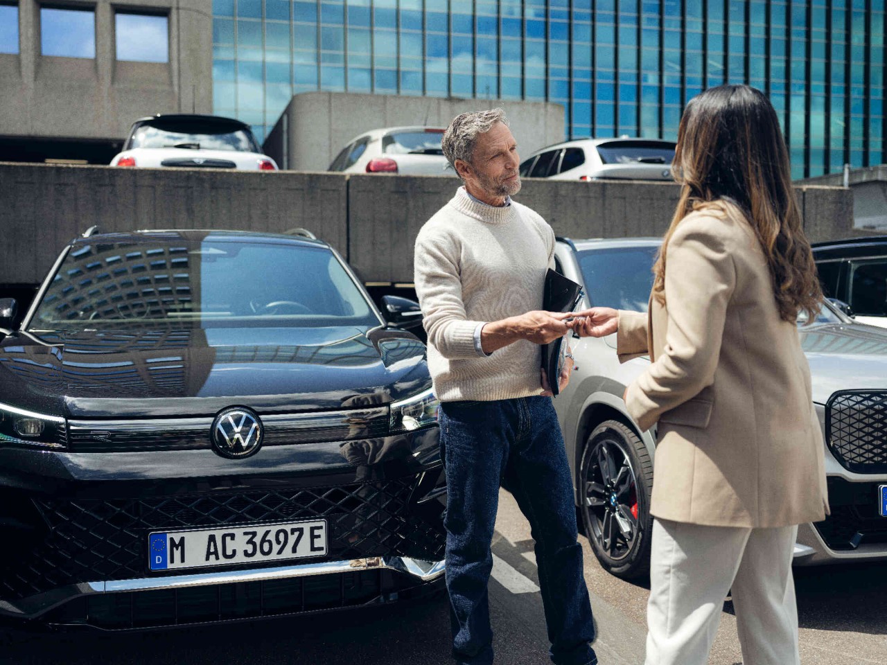 Mann ueberreicht Autoschluessel an Frau auf Parkplatz vor Buerogebaeude.