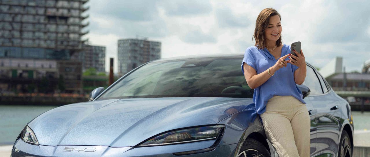 Eine Frau lehnt sich entspannt an ein blaugraues BYD Auto und nutzt ihr Handy w?hrend im Hintergrund ein Fluss und B?rogeb?ude zu sehen sind.