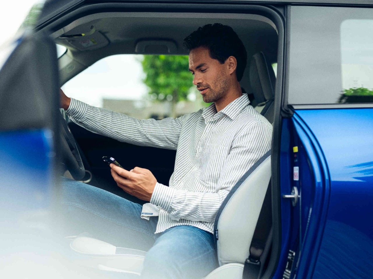 Mann sitzt auf Fahrersitz eines stehendes Autos schaut auf sein Handy Fahrertuer steht offen.