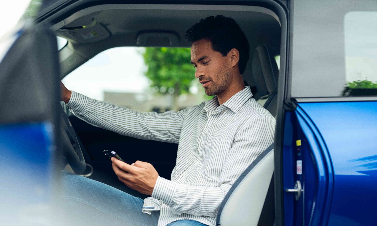 Mann sitzt auf Fahrersitz eines stehendes Autos schaut auf sein Handy Fahrertuer steht offen.