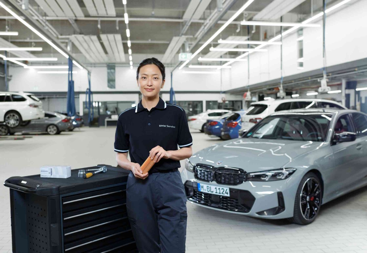 Frau steht neben einem Werkzeugwagen in einer Werkstattumgebung mit Autos im Hintergrund 