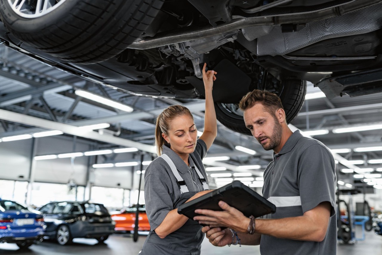 Frau zeigt mit dem Finger nach oben auf den Unterboden eines Fahrzeugs und schaut mit einem Mann auf ein Geraet 