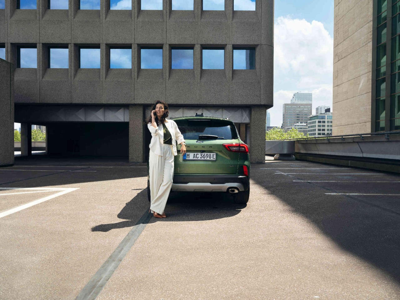 Frau lehnt an gruenen Ford auf Parkplatz vor modernen Gebaeuden.