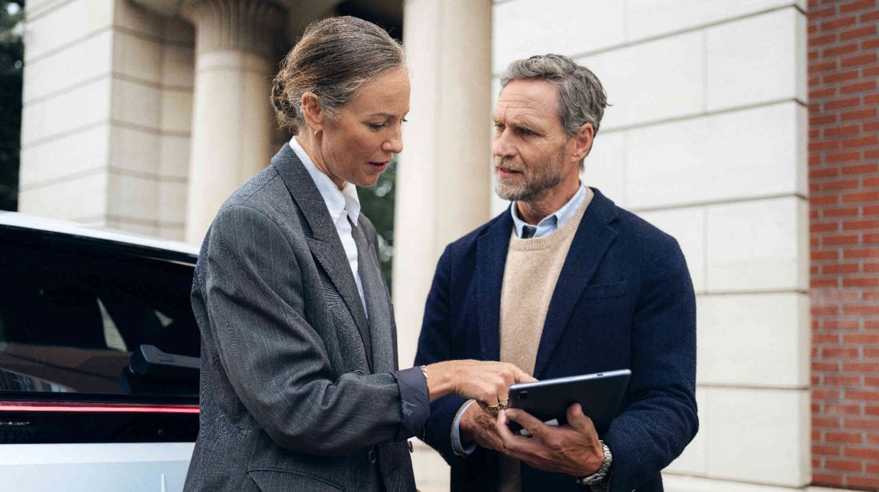 Frau unterhaelt sich mit Mann und zeigt auf Tablet in seiner Hand.