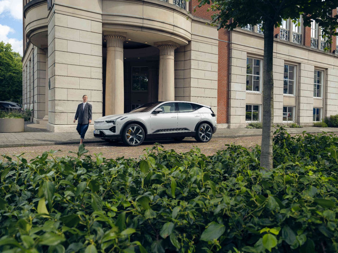 Frau geht vor einem geparkten silbernen Auto vor einem Gebaeude mit Saeulen.
