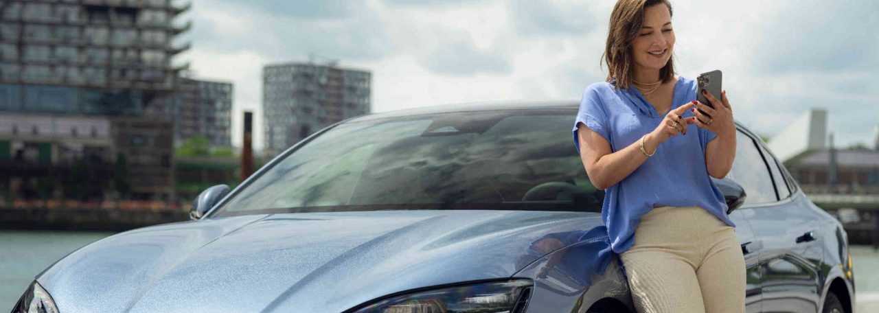 Eine Frau lehnt sich entspannt an ein blaugraues BYD Auto und nutzt ihr Handy w?hrend im Hintergrund ein Fluss und B?rogeb?ude zu sehen sind.