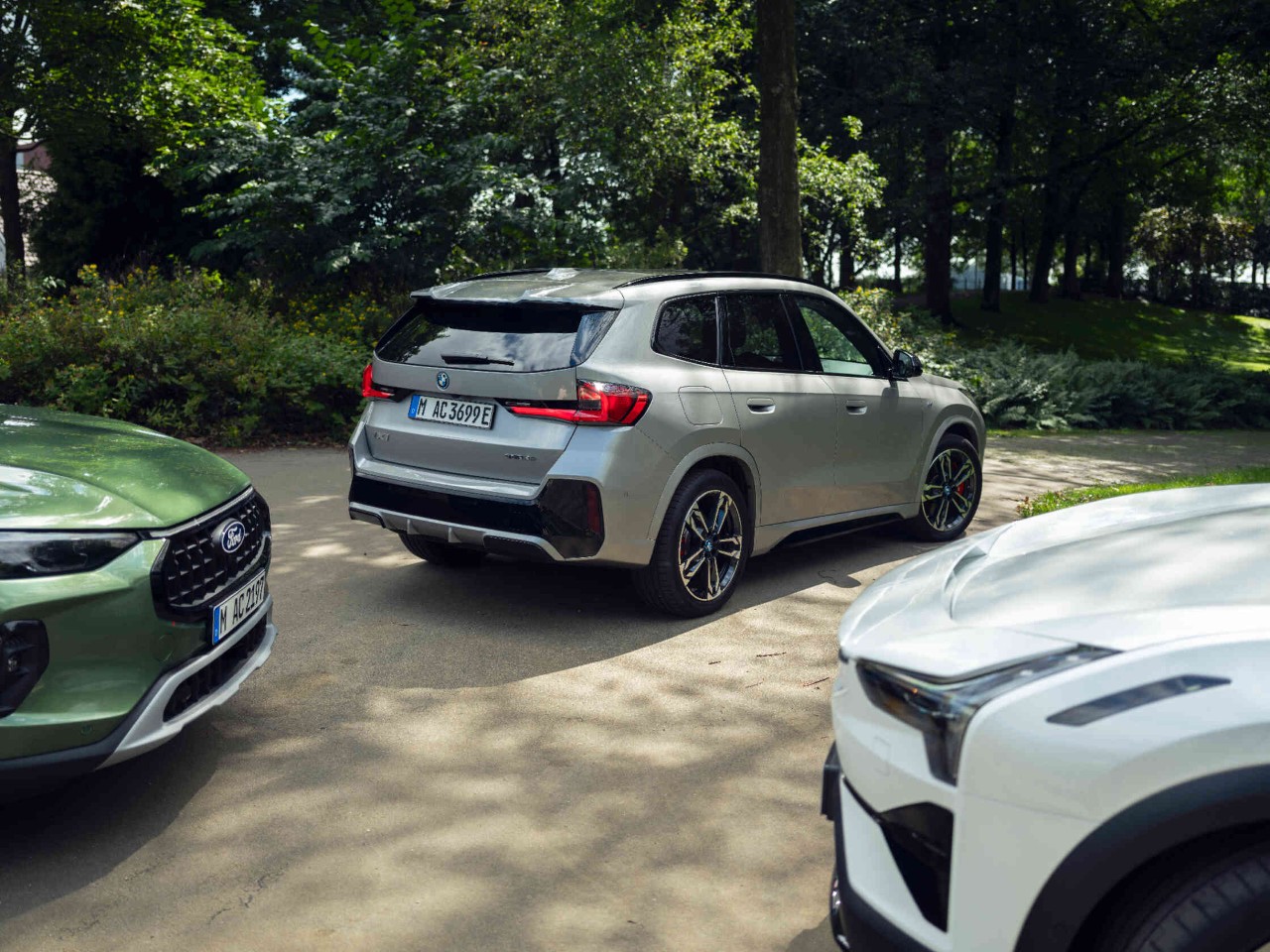 Drei Autos parken auf Parkplatz mit gruener Szenerie im Hintergrund.