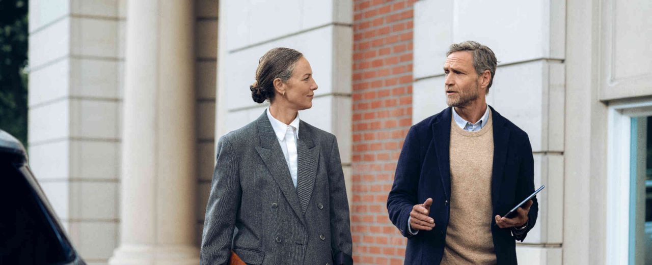 Frau mit Aktentasche geht und unterhaelt sich mit Mann mit Tablet vor elegantem Gebaeude.