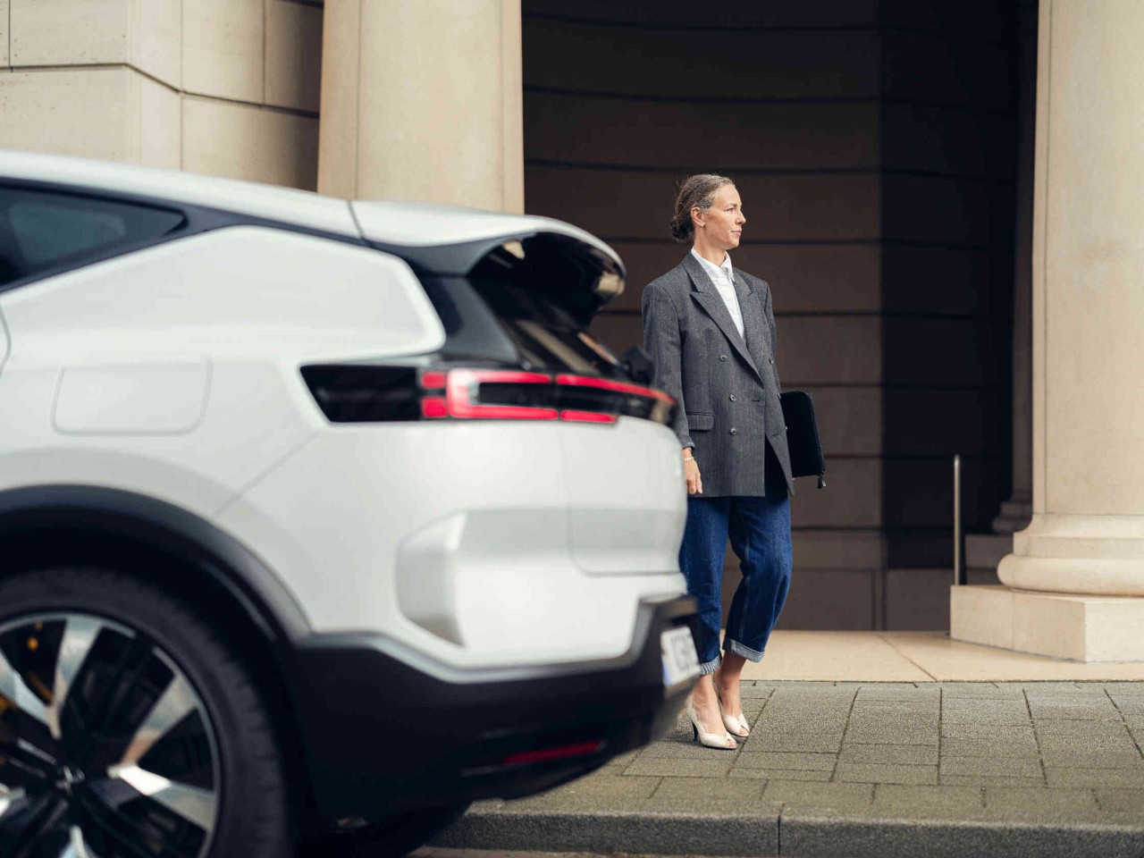 Frau steht auf dem Gehweg vor einem elegantem Gebaeude neben weissem Auto.