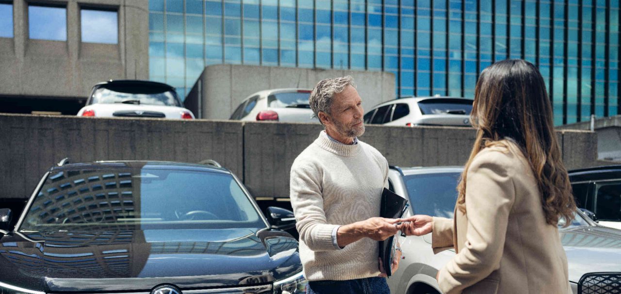 Mann ueberreicht Autoschluessel an Frau auf Parkplatz vor Buerogebaeude.