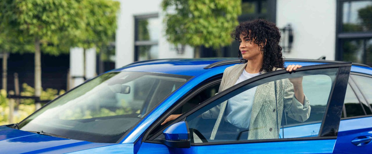 Frau steigt aus einem blauen Auto vor einem modernen Gebaeude aus.