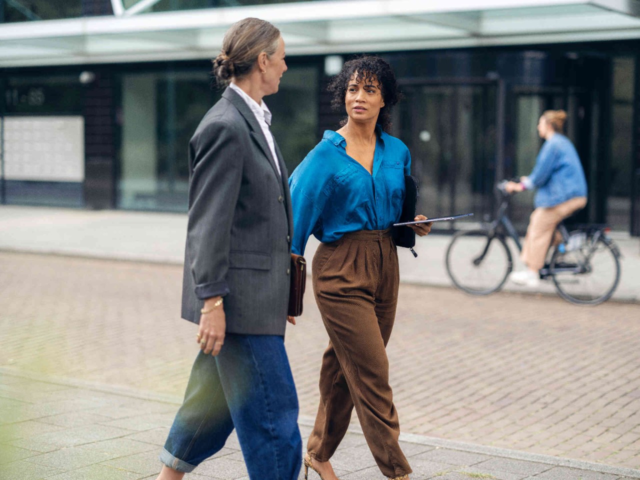 Zwei Frauen gehen auf Gehweg vor Gebaeude mit grossen Glasfenstern.