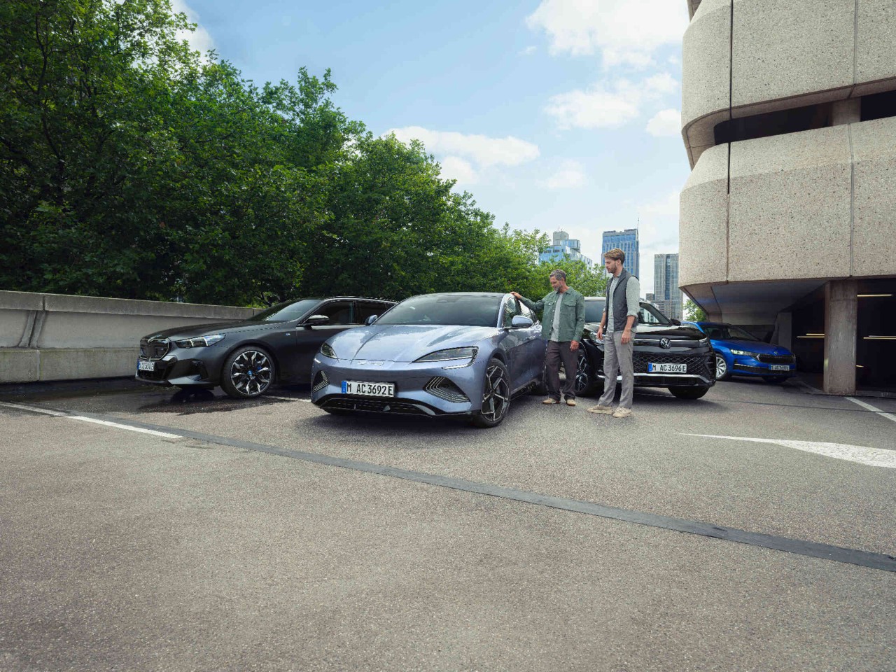 Zwei Maenner neben drei Autos auf Parkplatz vor Parkdeck.