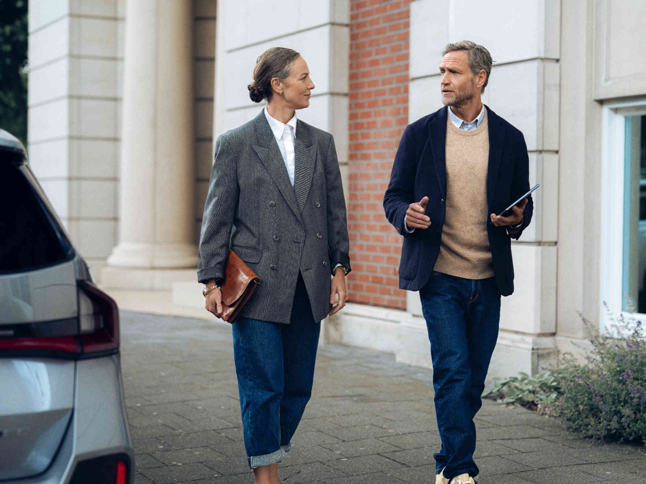 Frau mit Aktentasche geht und unterhaelt sich mit Mann mit Tablet vor elegantem Gebaeude.