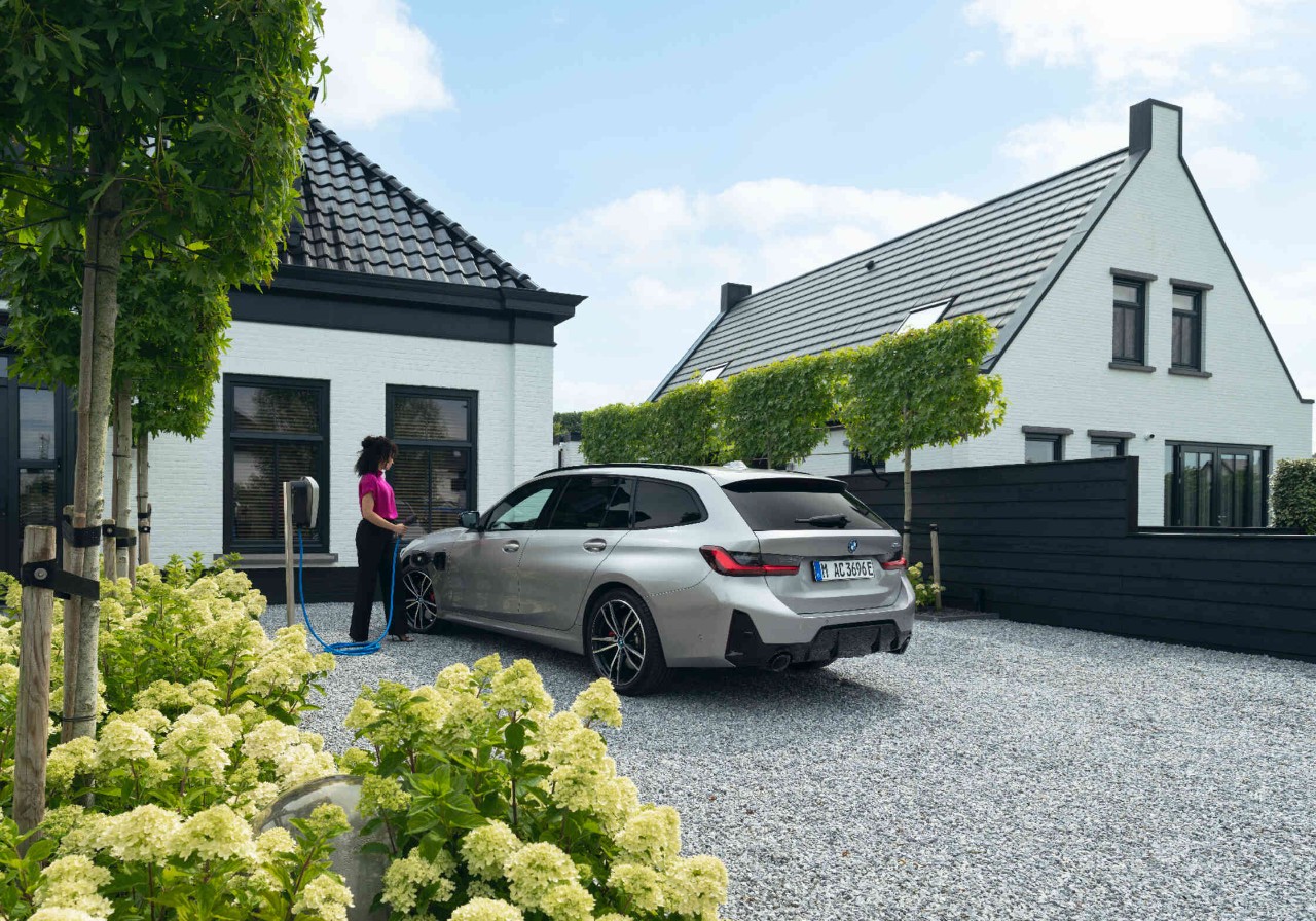 Frau steht in Einfahrt neben einem grauen BMW Auto und steckt einen Stecker in den Ladenanschluss.