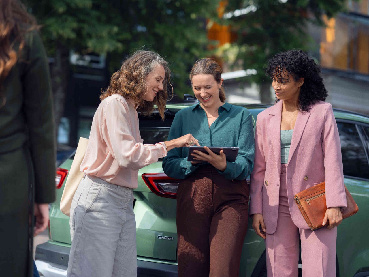 Drei Frauen diskutieren vor einem gruenem Auto mit Tablet in der Hand.