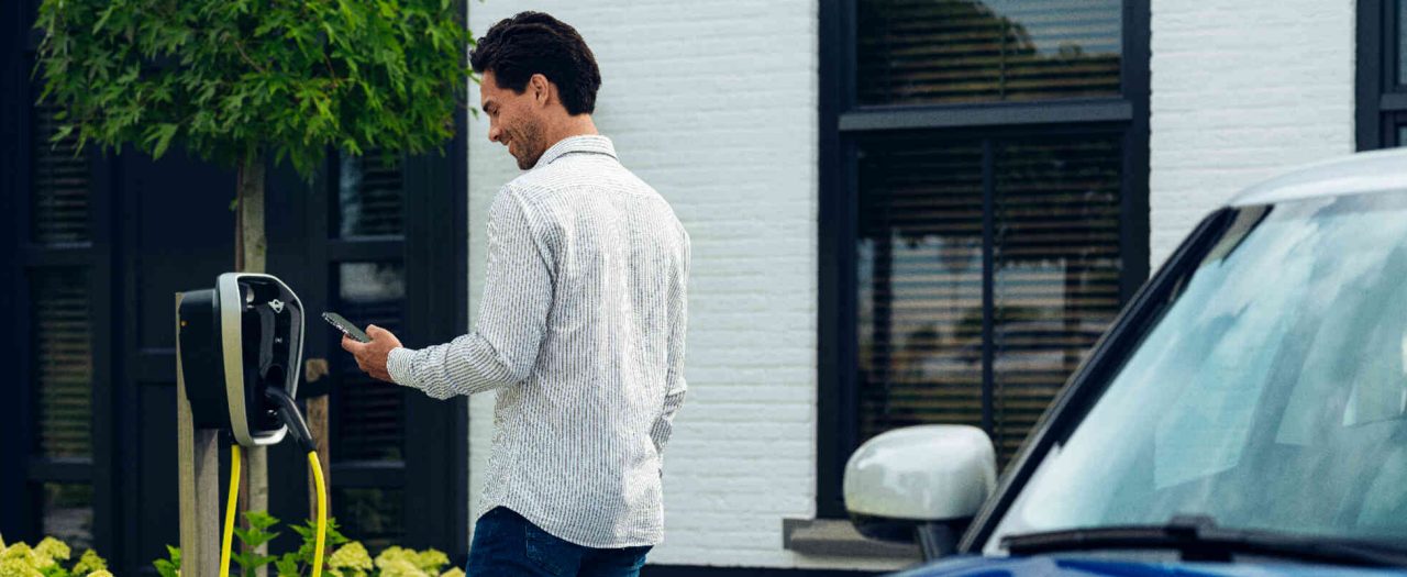 Mann schaut auf Handy und steht vor einer Heimladestation hinter ihm blauer MINI.