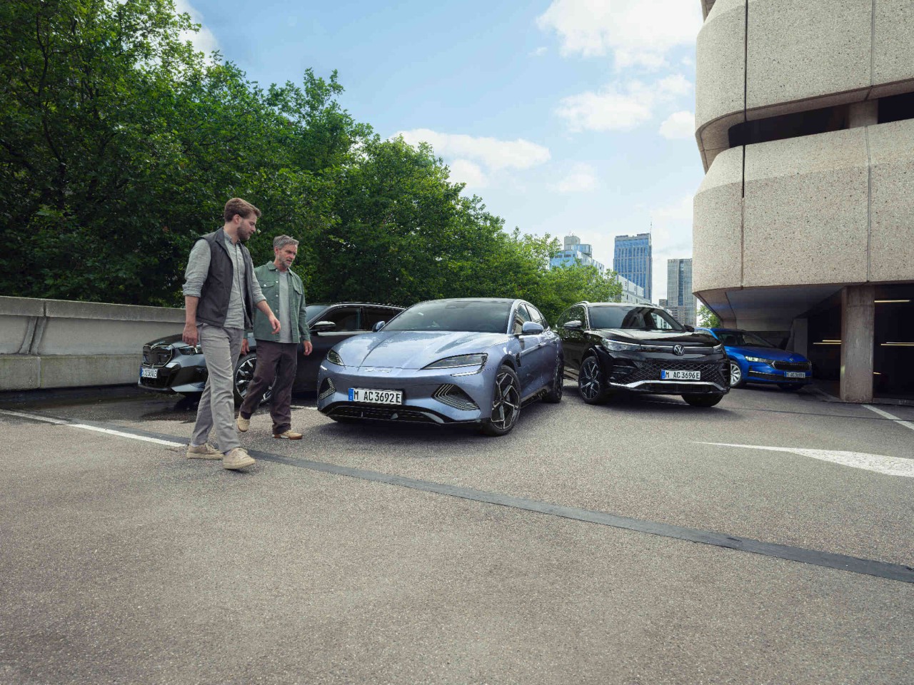 Zwei Maenner vor drei Autos auf Parkplatz vor Parkdeck.