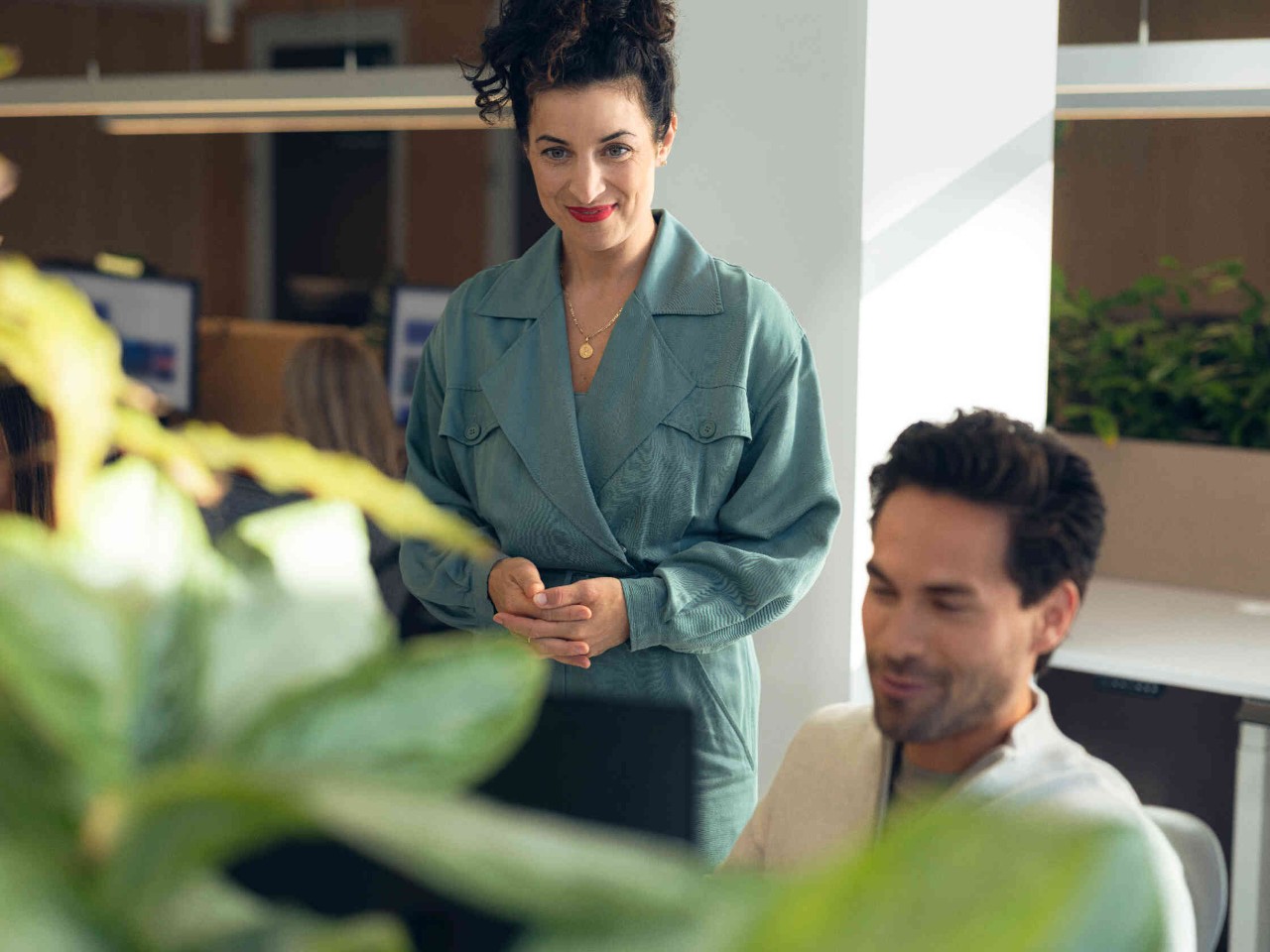 Zwei Personen blicken laechelnd auf Computer in hellen Buero mit Pflanzen.