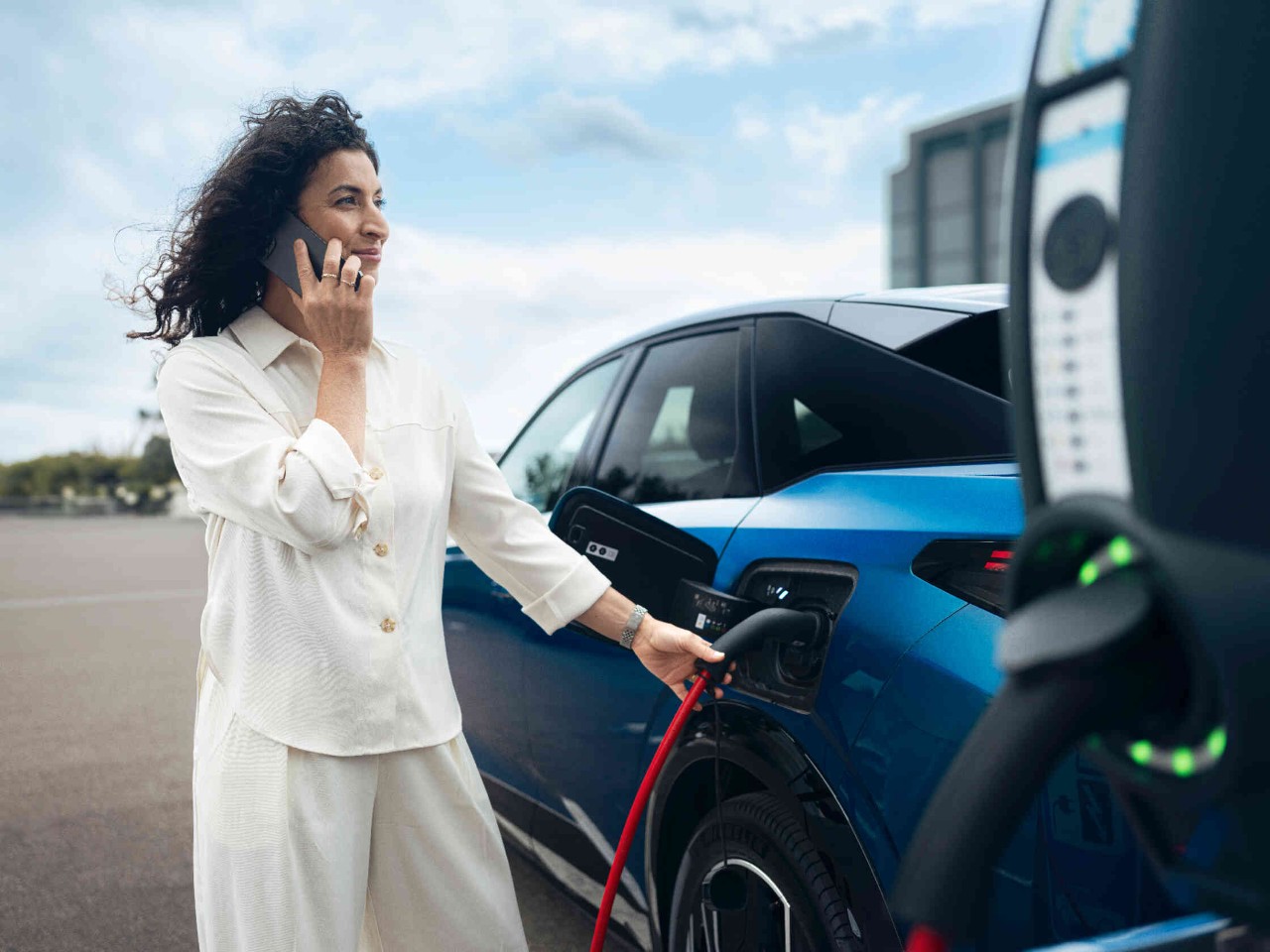 Frau in cremefarbenem Outfit laedt blaues Elektroauto an Ladestation auf.