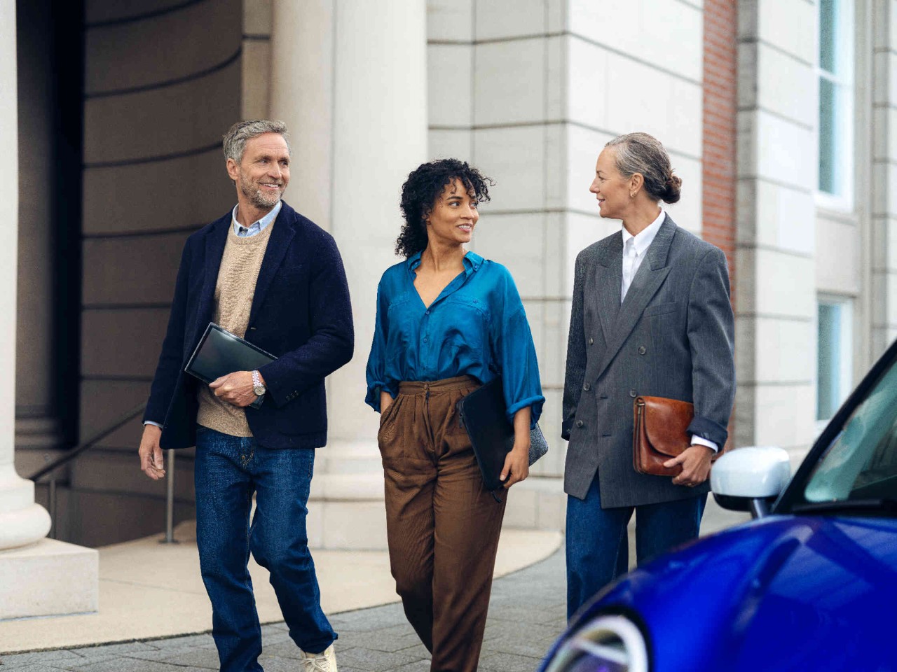 Drei Personen gehen auf einem Gehweg vor einem elegantem Gebaeude.