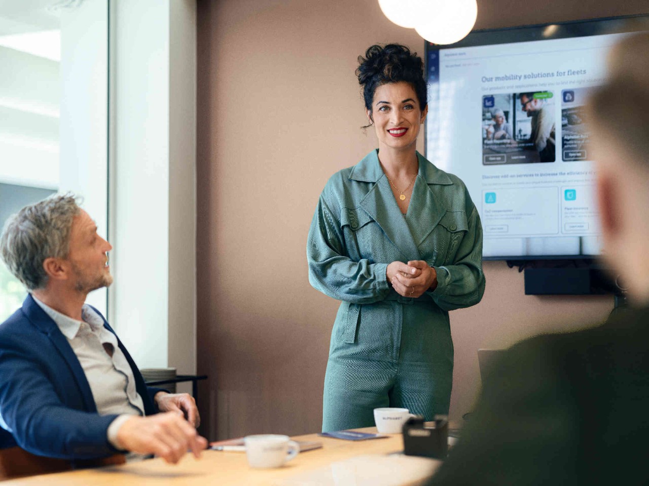 Frau in gruenem Blazer praesentiert vor Bildschirm im Konferenzraum.