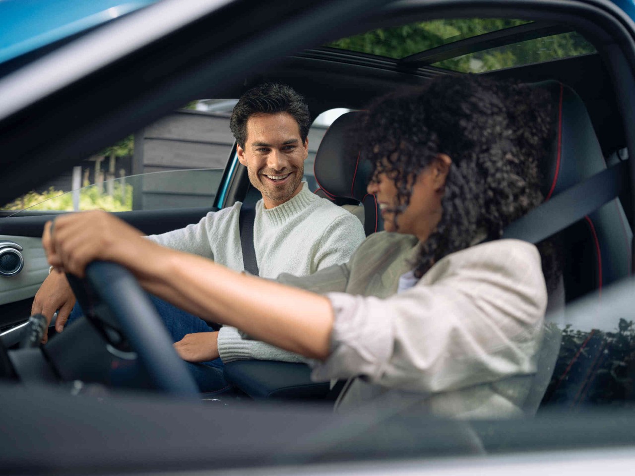 Zwei Personen sitzen in einem modernen Auto mit Panoramadach die Fahrerin haelt das Lenkrad.