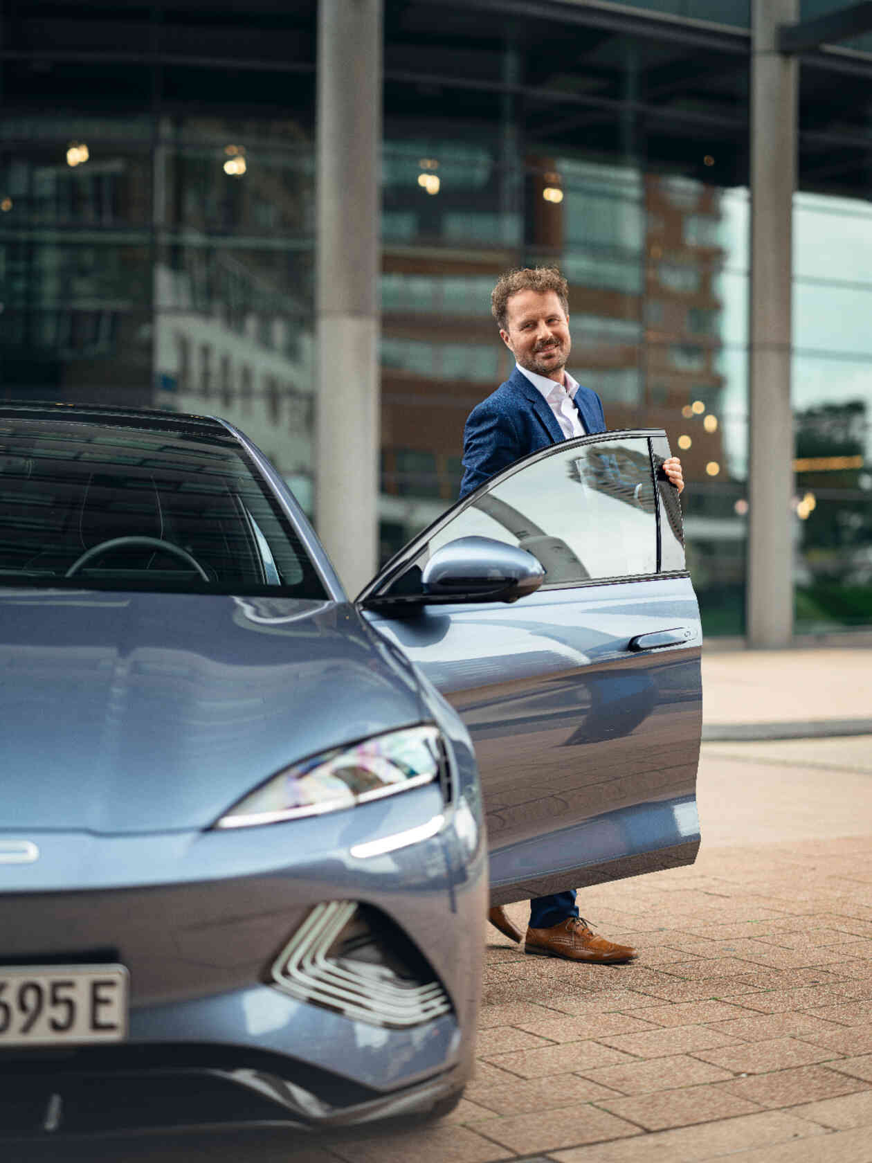 Mann steigt aus blaugrauen Auto aus und laechelt in Richtung Kamera.