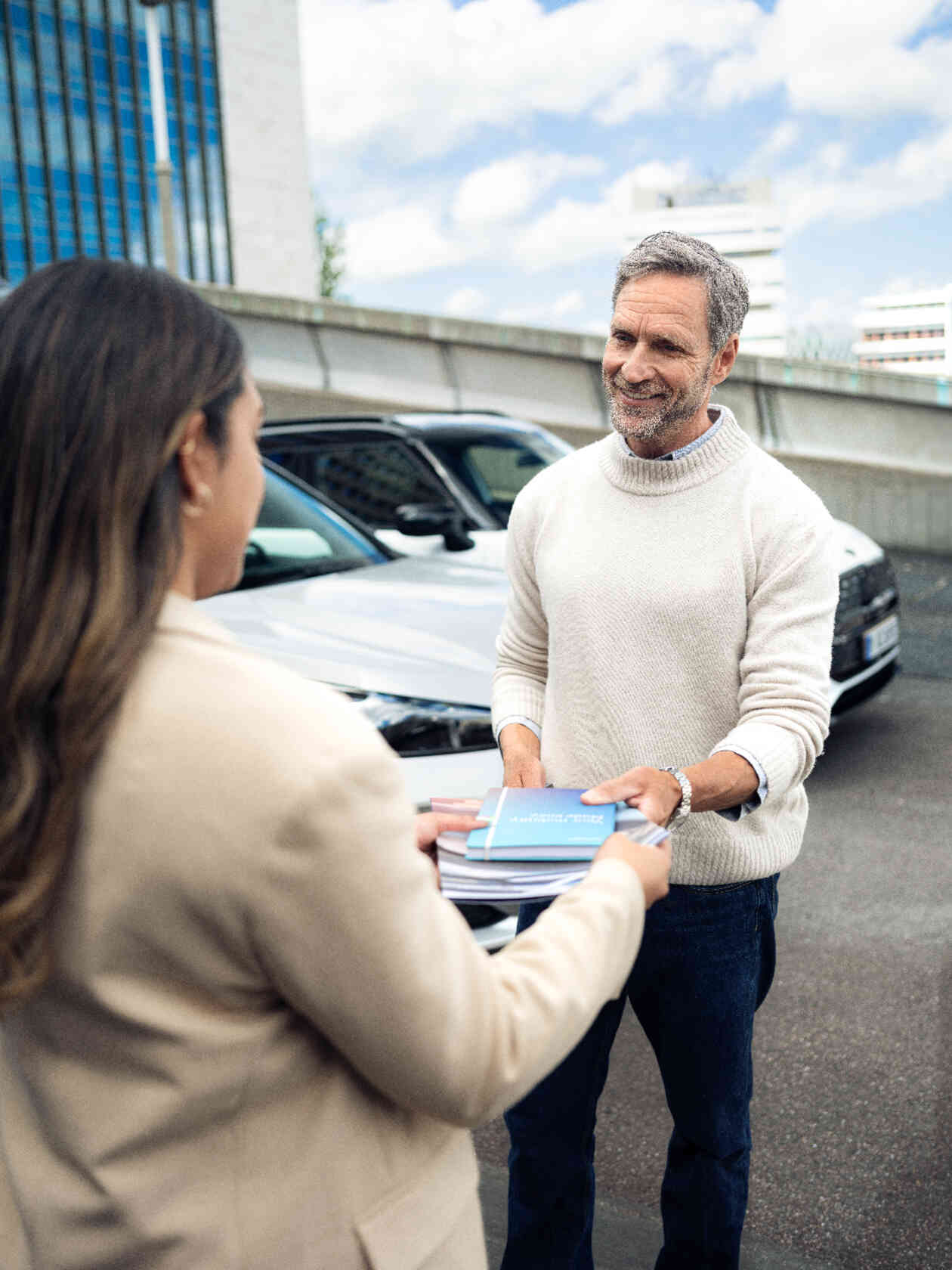 Frau ueberreicht Dokumente an Mann auf Parkplatz.