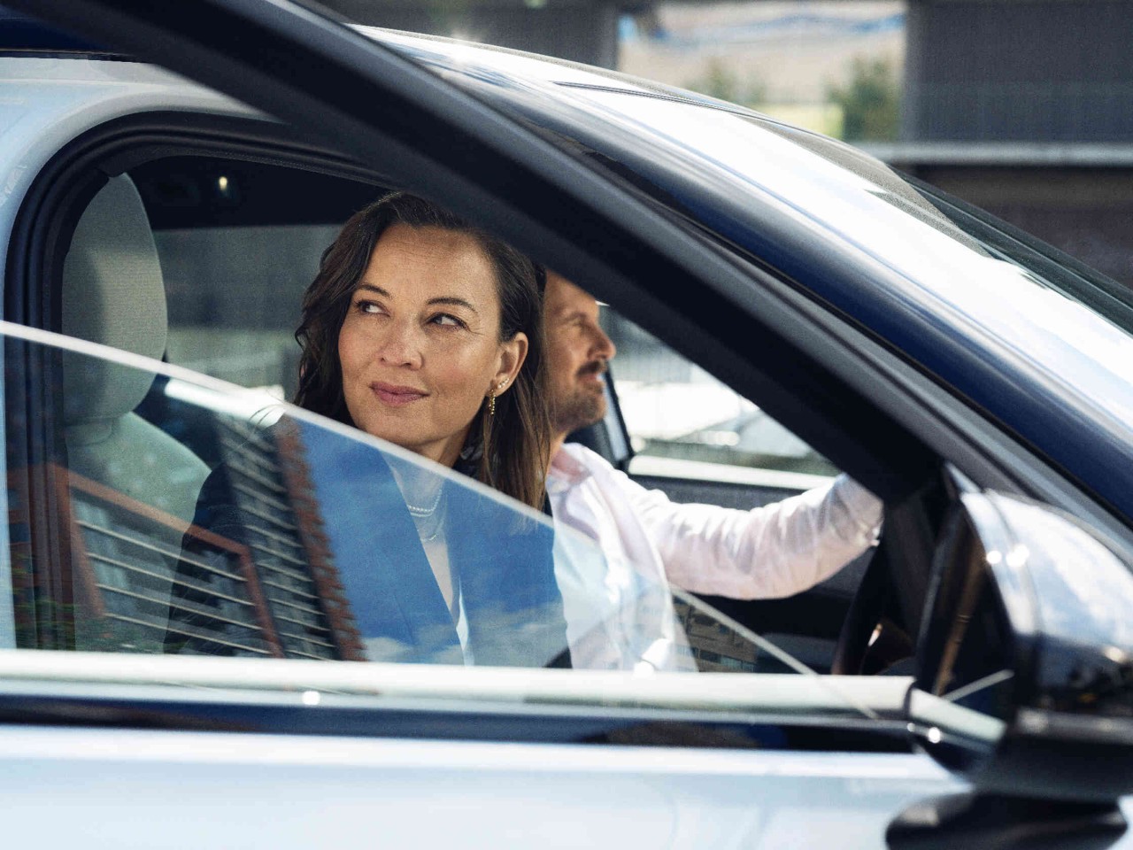 Ein Mann sitzt am Steuer eines Autos während eine Frau auf dem Beifahrersitz einsteigt oder aussteigt und sich umschaut.