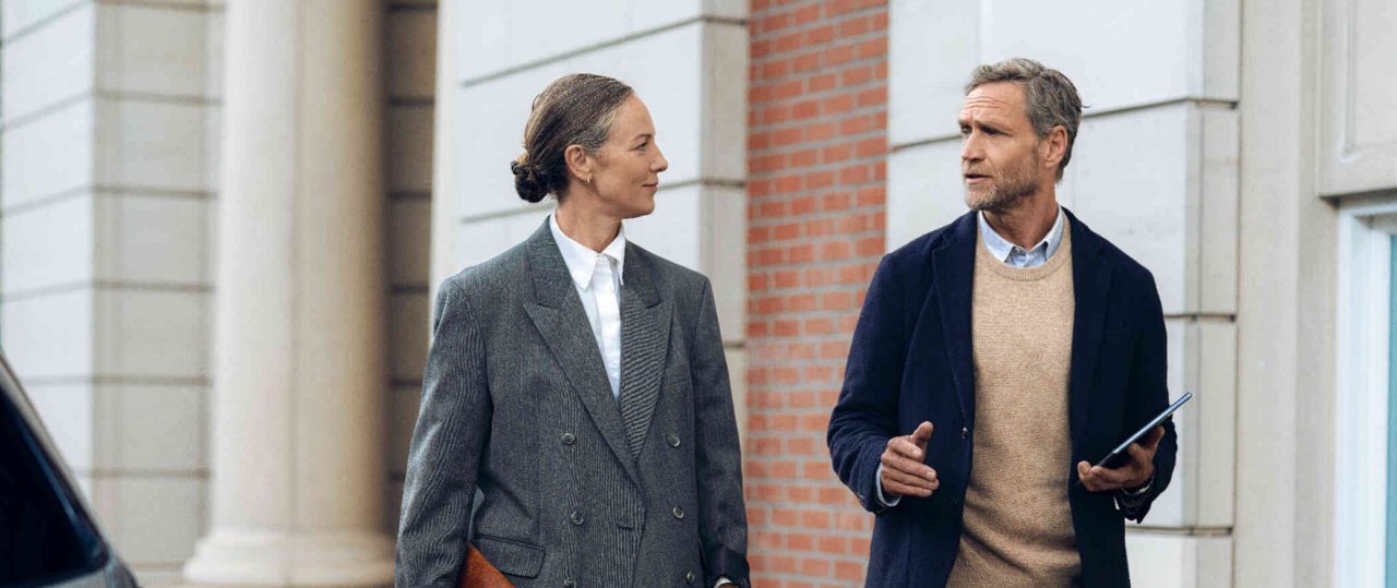 Frau mit Aktentasche geht und unterhaelt sich mit Mann mit Tablet vor elegantem Gebaeude.