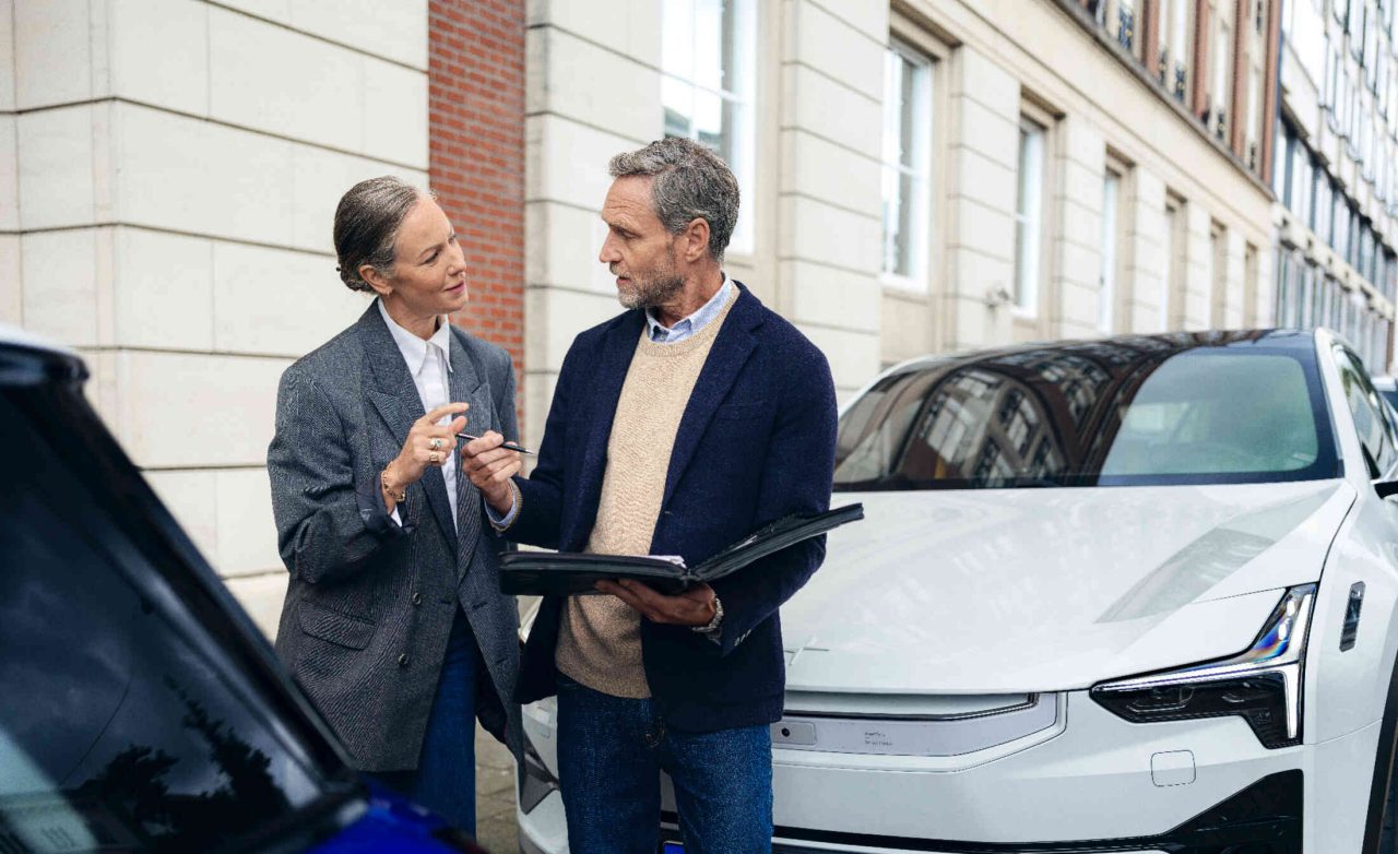 Frau und Mann unterhalten sich zwischen zwei geparkten Autos in urbanem Umfeld.