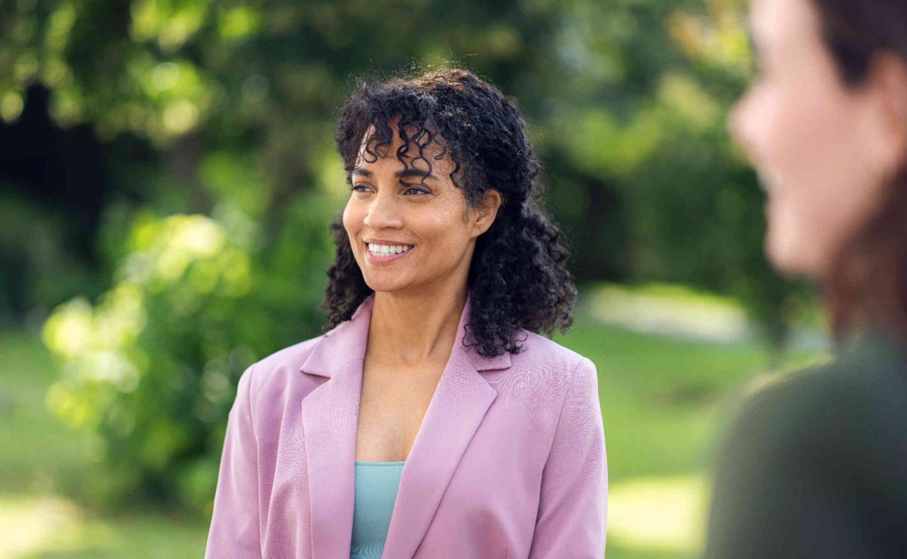 Frau in rosa Blazer und brauner Tasche steht in gruenem Park und laechelt.