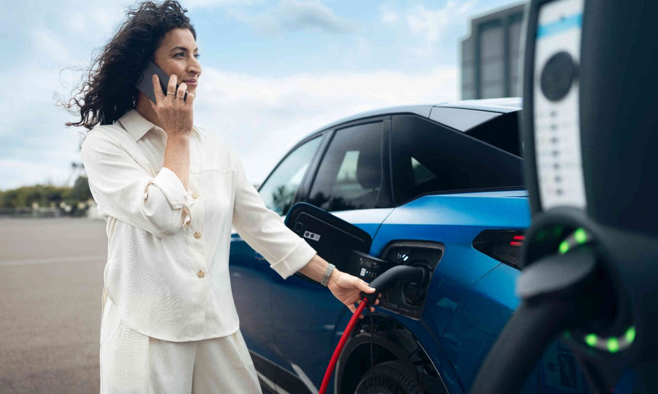 Frau in cremefarbenem Outfit laedt blaues Elektroauto an Ladestation auf.