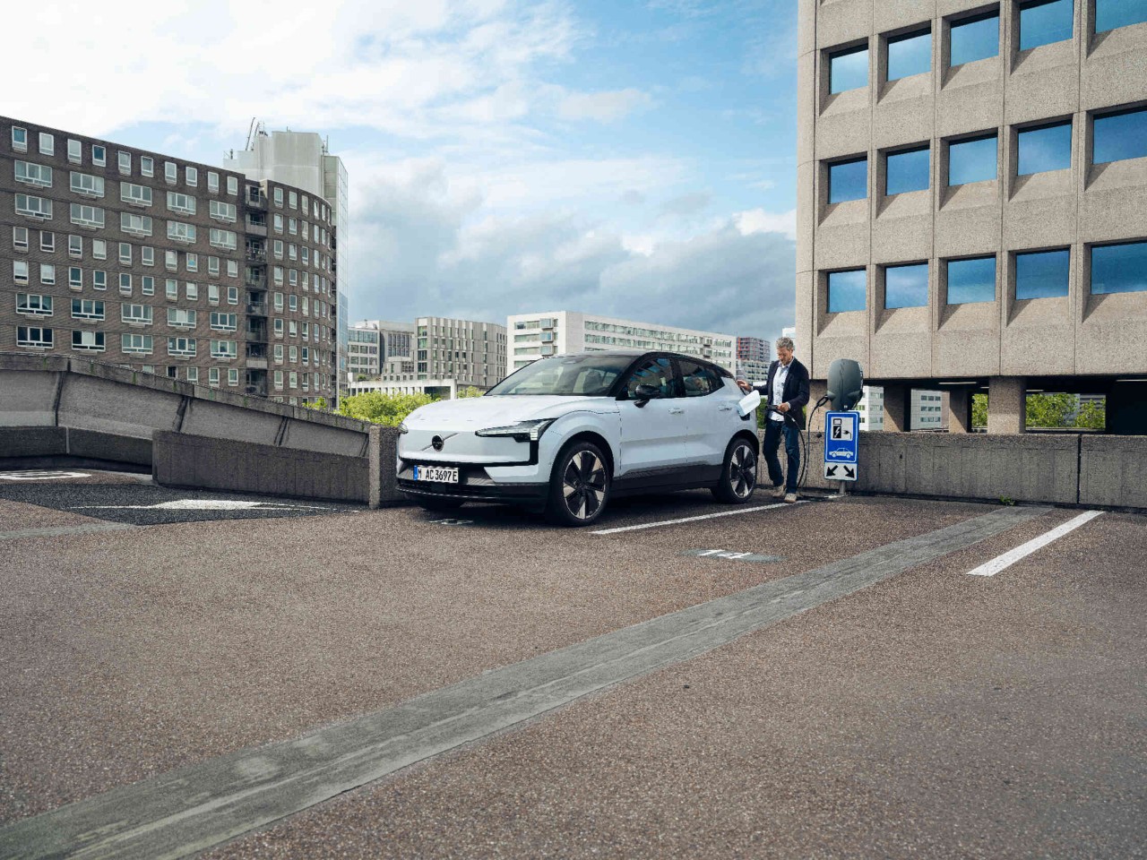 Mann laedt Elektroauto auf staedtischem Parkdeck mit modernen Gebaeuden.