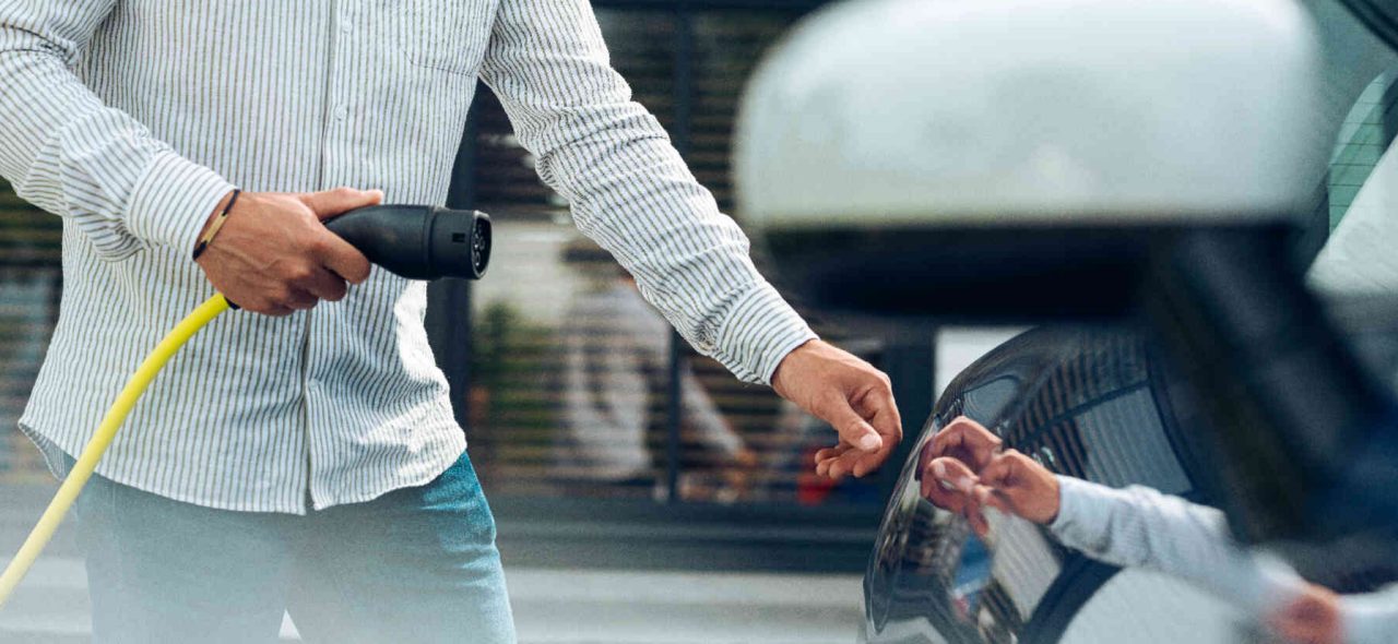 Mann haelt Stecker einer E-Ladestation in der Hand und oeffnet die Klappe zum Ladeanschluss eines Autos.