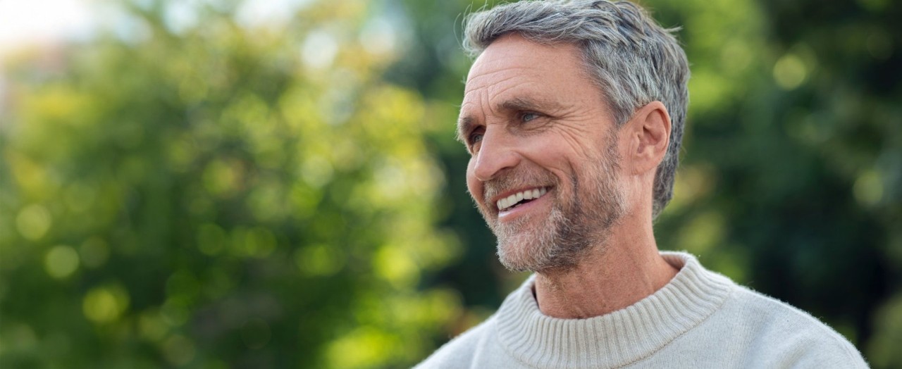 Mann mit grauem Haar und hellem Pullover steht im Freien und laechelt.