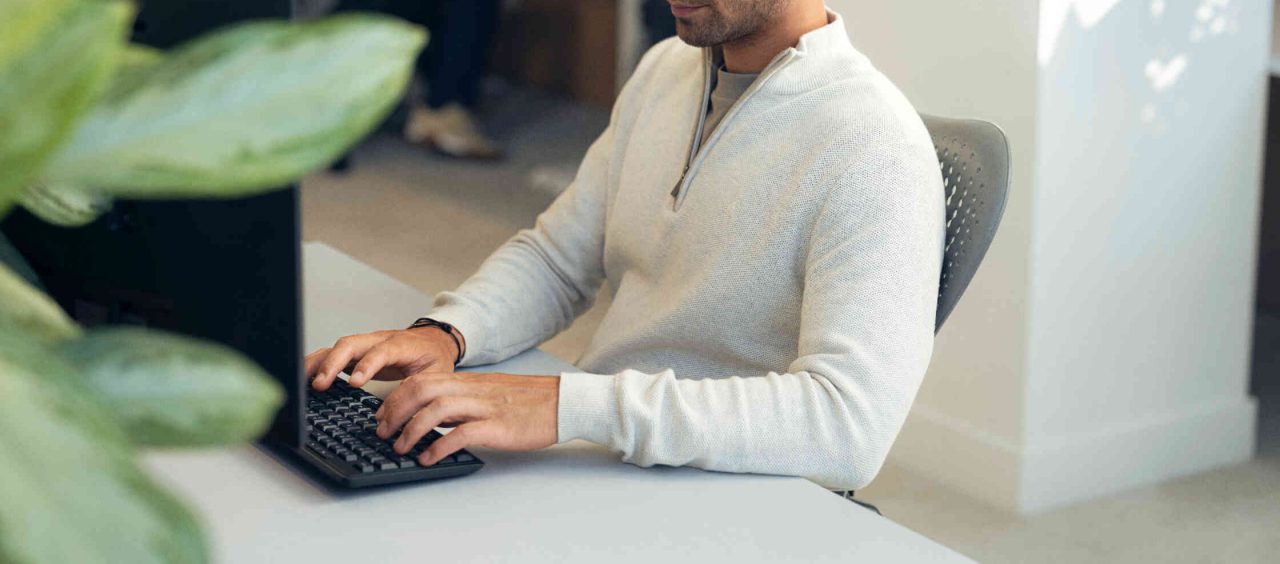 Mann tippt auf Tastatur vor Computer in hellen Buero mit Pflanzen