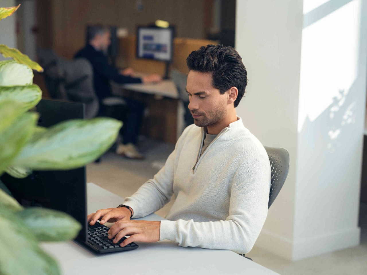 Mann tippt auf Tastatur vor Computer in hellen Buero mit Pflanzen