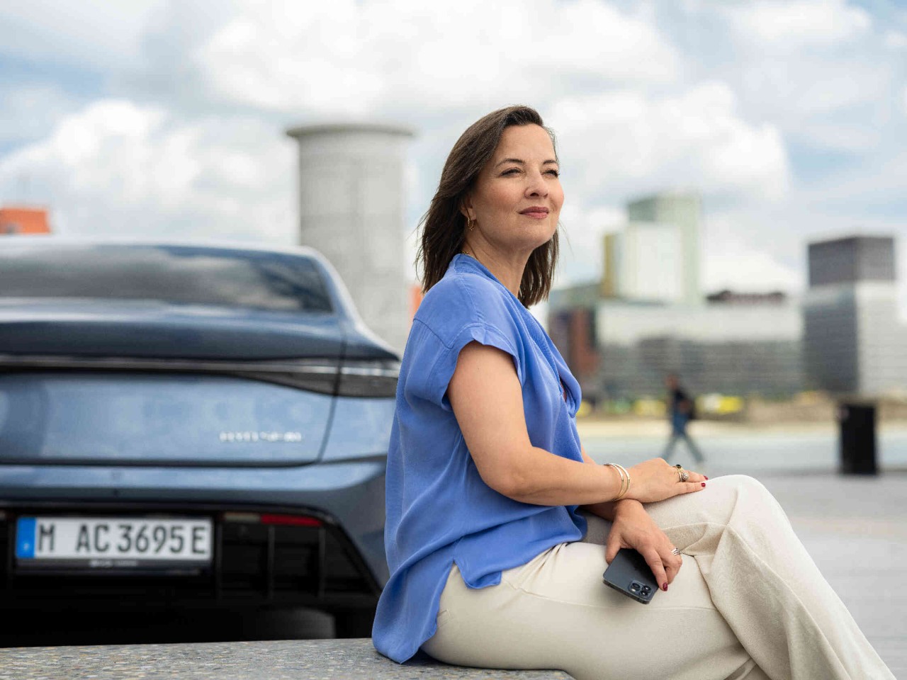 A woman sits on a bench and gazes into the distance while a car and modern buildings are visible in the background.